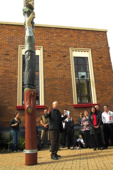 Picture of David Sarich standing next to the Dalmatian Pou, which he carved, explaining the the pou's design at its unveiling ceremony.