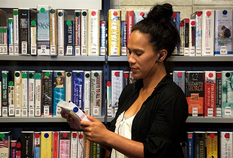 Picture of a woman listening to a talking book among library shelves of talking books. 