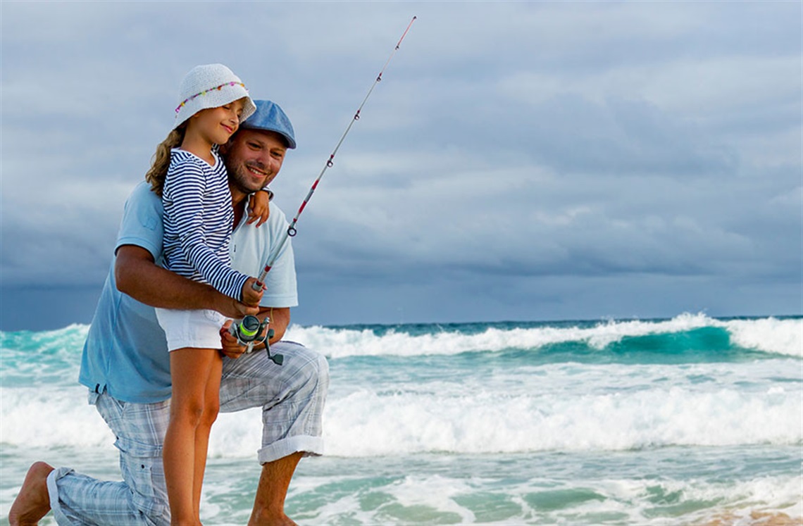 A man and a young girl holding a fishing rod in front of the ocean. 