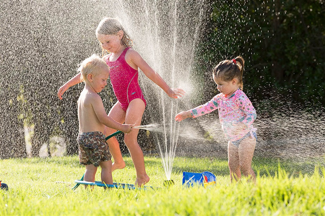 A young girl and a boy and girl toddler play outdoors in their togs with a hose and sprinklers on the grass. 