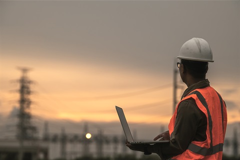 A man in high-vis vest holds a laptop and looks out over a power gird at dusk. 