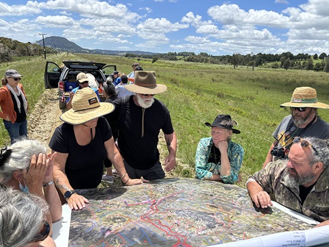 Several people gather around looking at a map on the bonnet of a car in a paddock. 