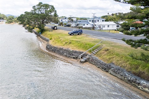 A photo of the Ngunguru seawall looking from the water back to houses and cars parked on the shore. 