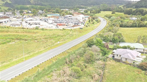 A view of Springs Flat with a road through the middle, a couple of houses on the right and a village in the background.