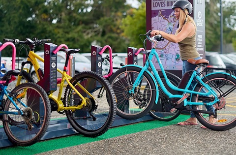 A young woman with long blond hair and wearing a cycle helmet places her bright blue bike into a Locky Dock parking station. 