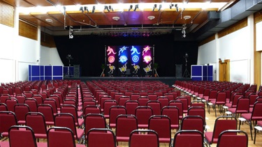 Behind angle of the Exhibition Hall theatre layout with chairs evenly spread out facing the stage.