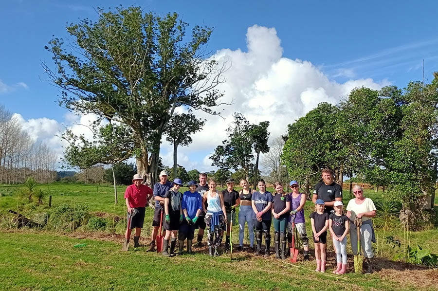 A group of people at a community planting day with trees behind them. 