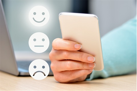 Person holding a mobile phone with laptop in background and 3 emoji faces for satisfaction rating. 