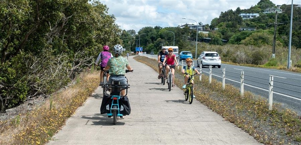 Photo showing a family of cyclists riding past a pair of commuting cyclists on the Onerahi Shared Path, separated from a busy road. 